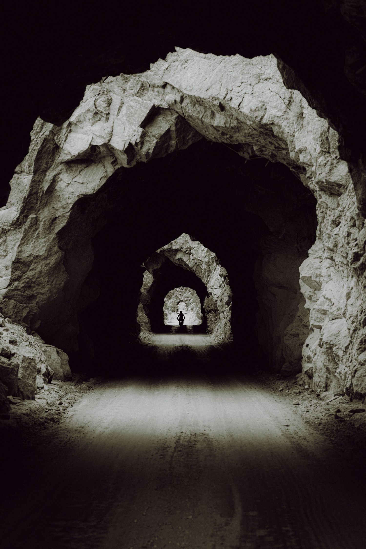 Silhouette of a person standing at the end of a dark rocky tunnel with multiple stone arches receding into the distance.