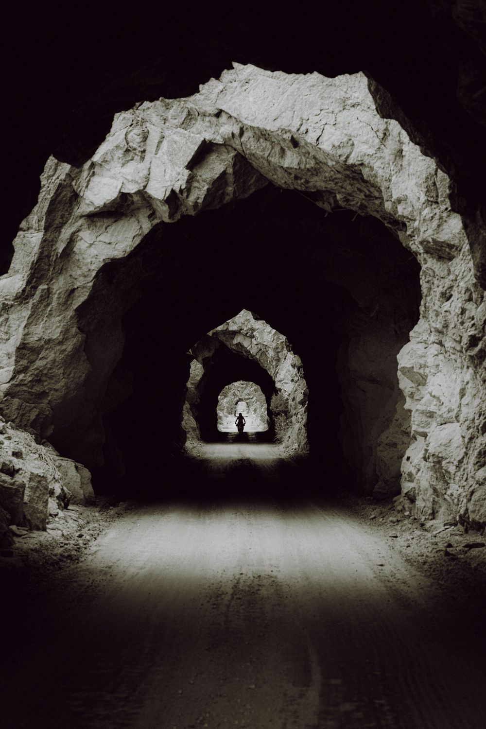 Silhouette of a person standing at the end of a dark rocky tunnel with multiple stone arches receding into the distance.