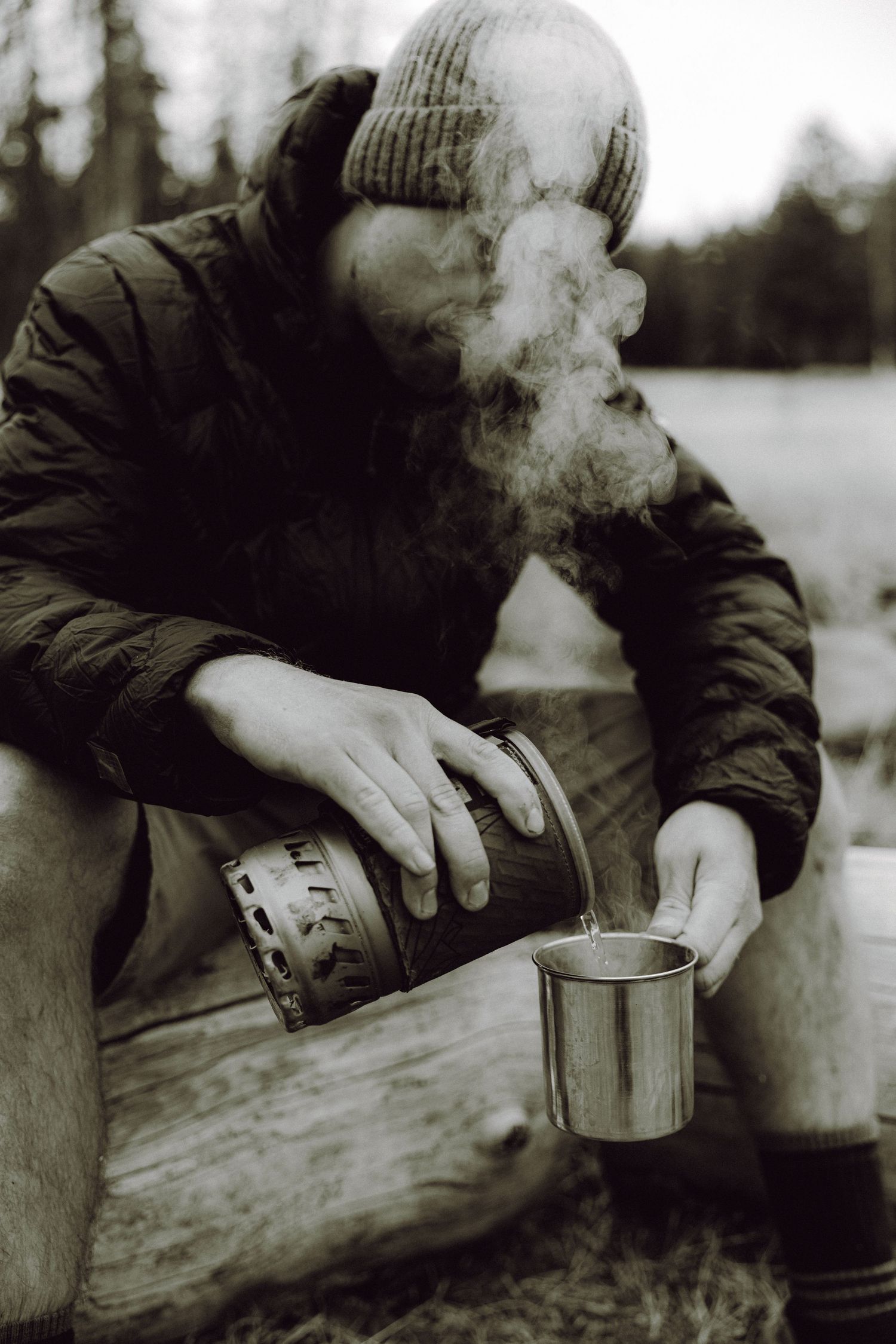 Bearded man in knit hat pouring hot steaming liquid into metal camping cup outdoors in winter forest setting.