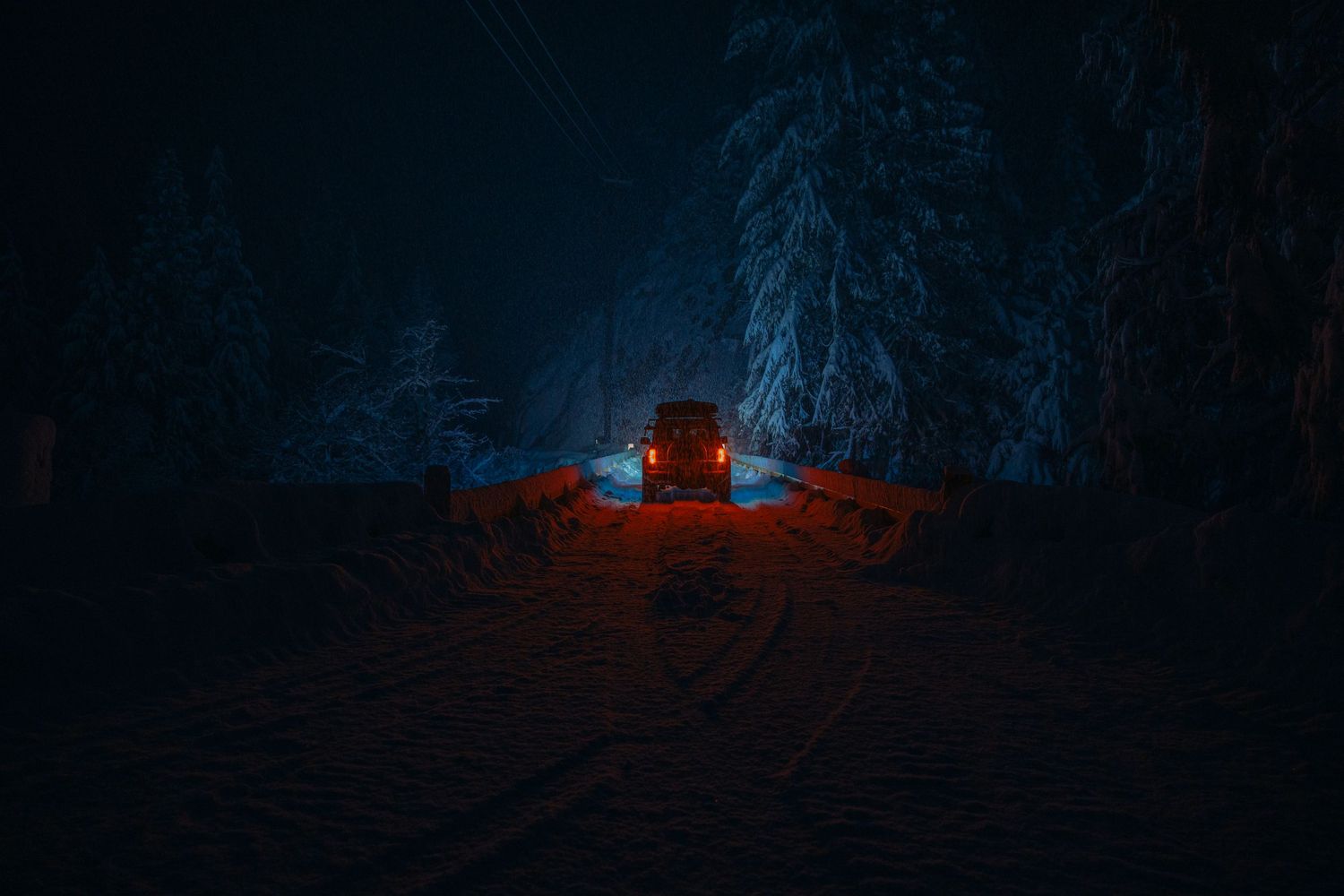 Silhouette of a Ford Bronco on a bridge during a snowy night. Taillights glowing red set against the cold night