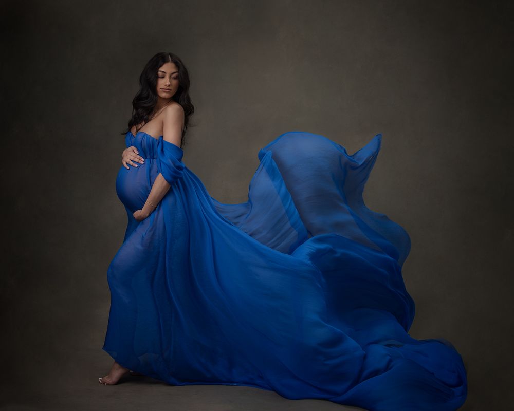 Dramatic maternity photo with flowing blue dress against dark background.