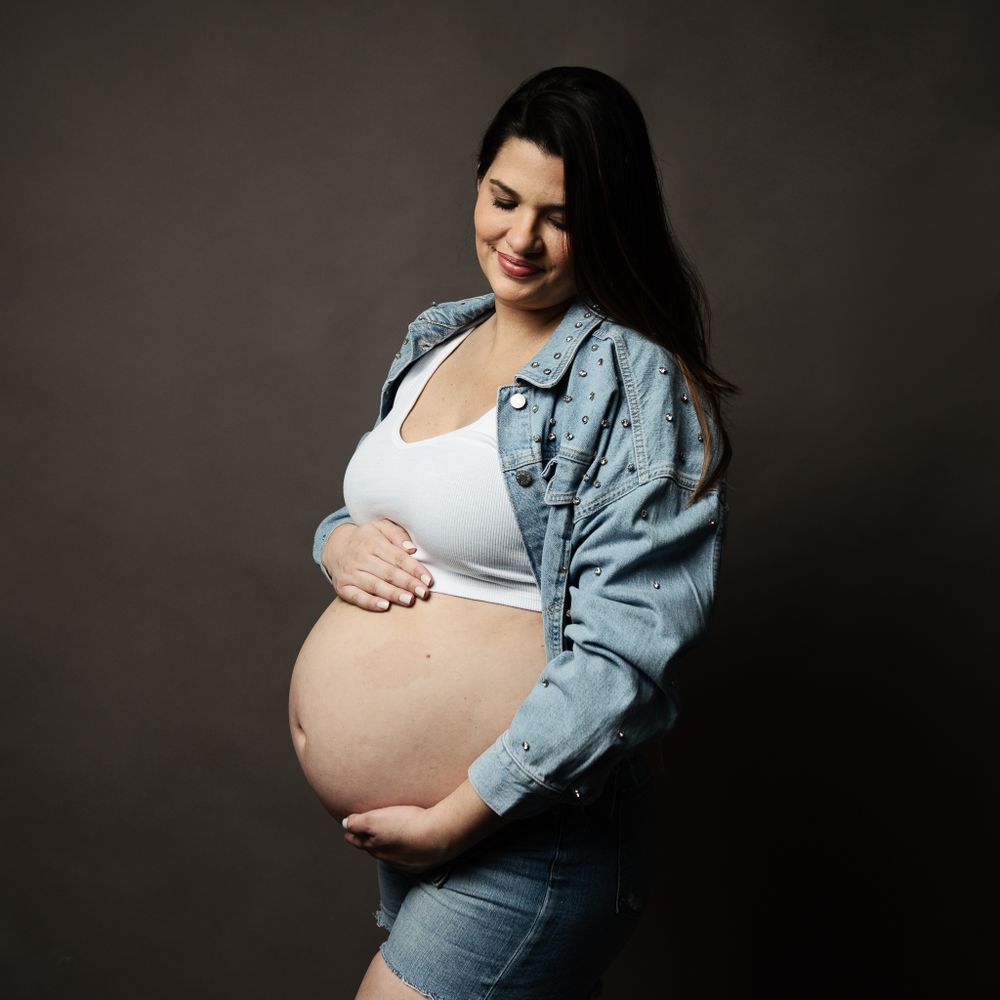 Maternity portrait featuring person in denim jacket and white top against dark background.