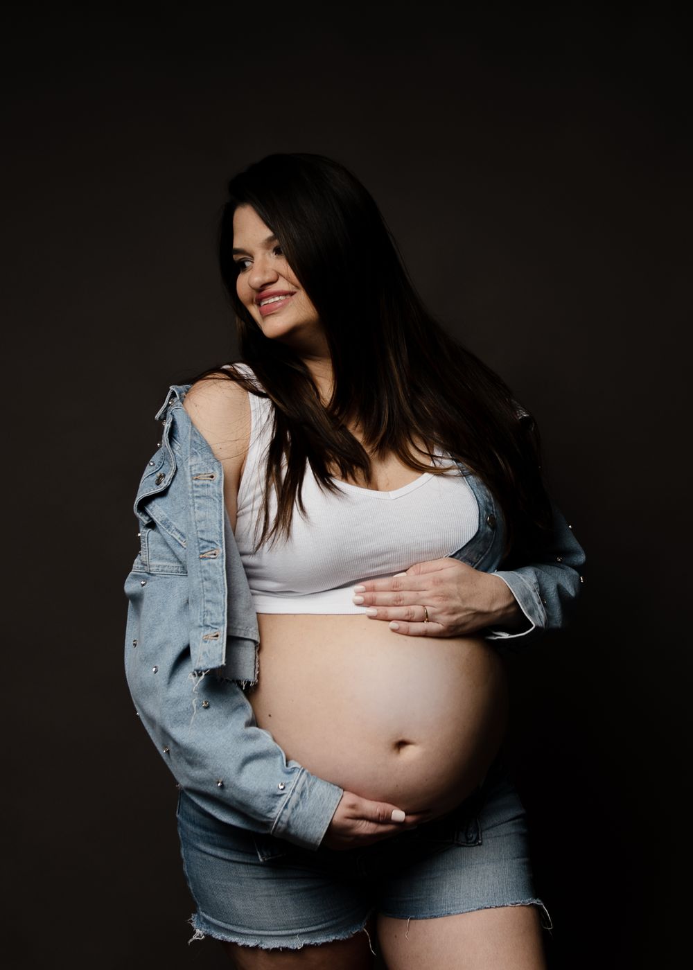 A casual maternity photo showing a woman in denim shorts and jacket against a dark background.