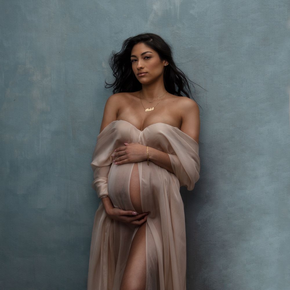 Artistic maternity portrait in sheer beige dress against textured wall.