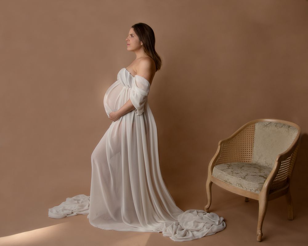 Elegant maternity portrait in flowing off-shoulder white gown beside vintage chair.