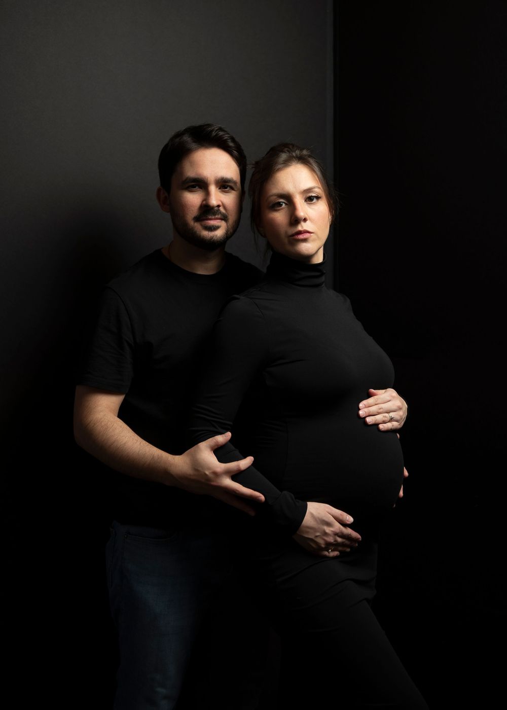 Dramatic low-key maternity portrait of couple in black clothing against dark background.