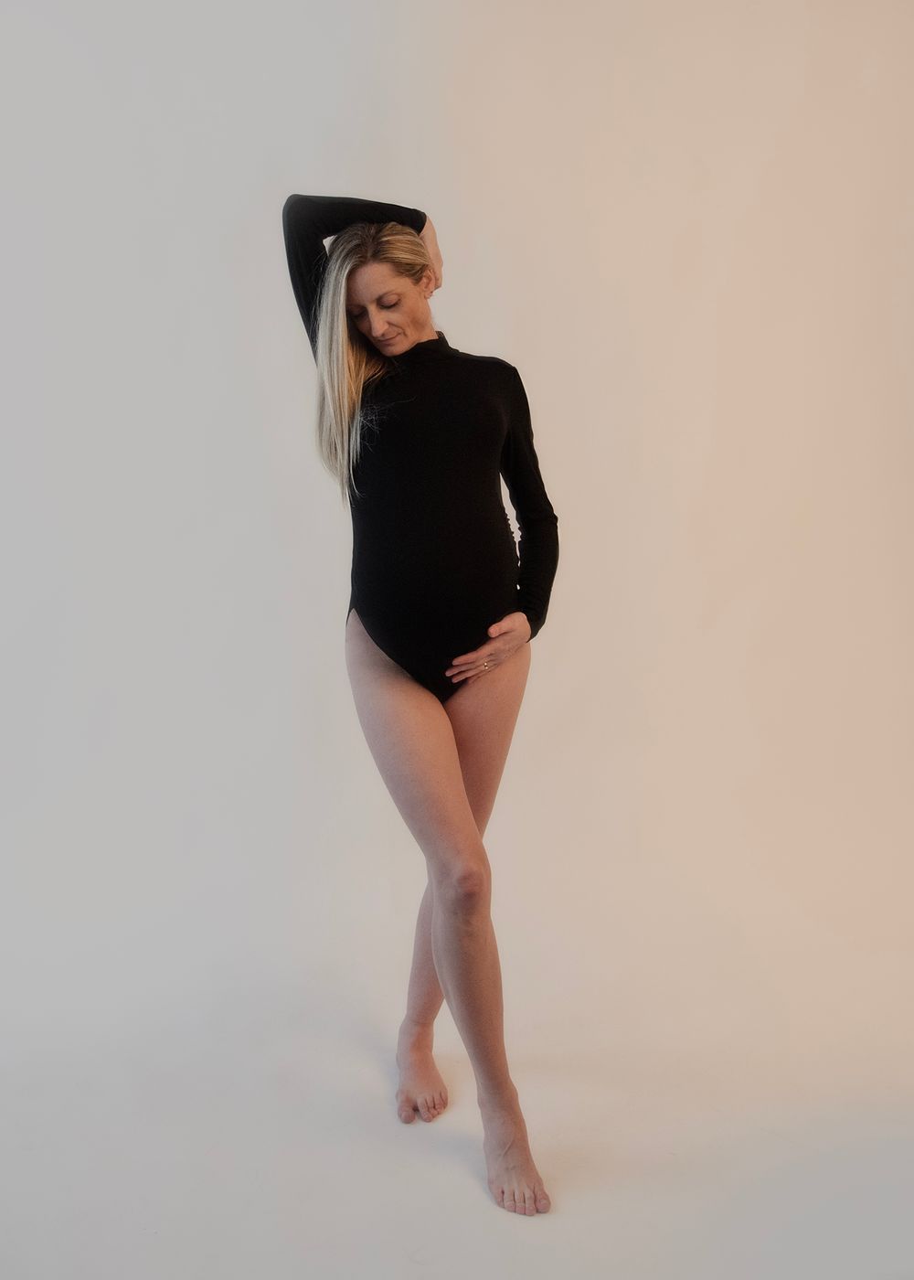 Artistic maternity pose in a black bodysuit against a light background.