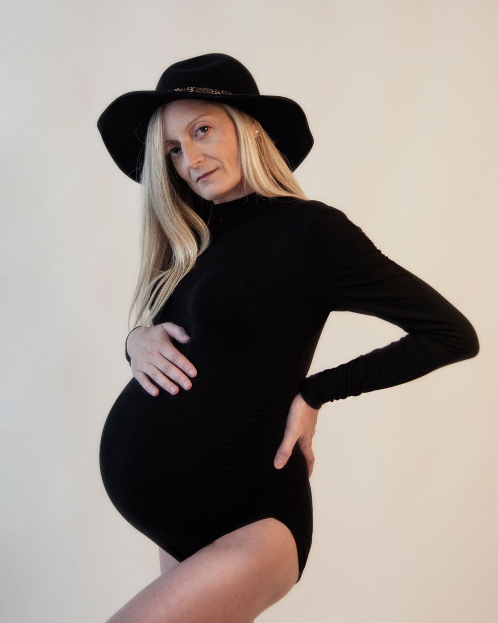 Stylish maternity portrait with black hat and fitted dress against white background.
