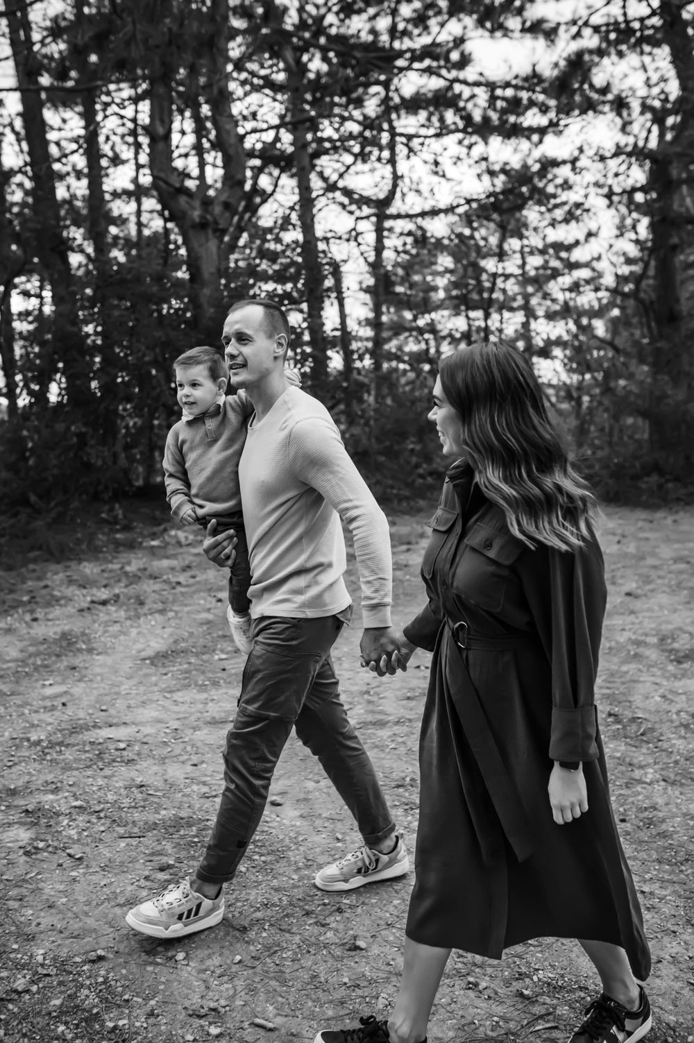 A family walks together on a forest trail in black and white, holding hands on an autumn afternoon.