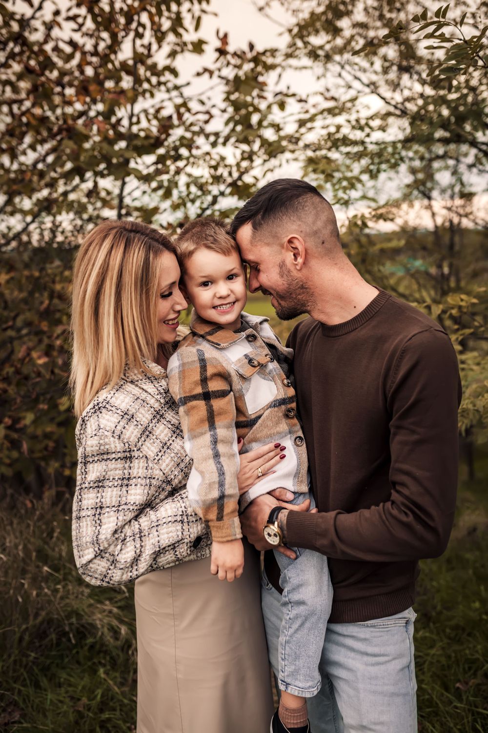 Adorable young family embracing outdoors in autumn with trees and soft natural lighting in the background.