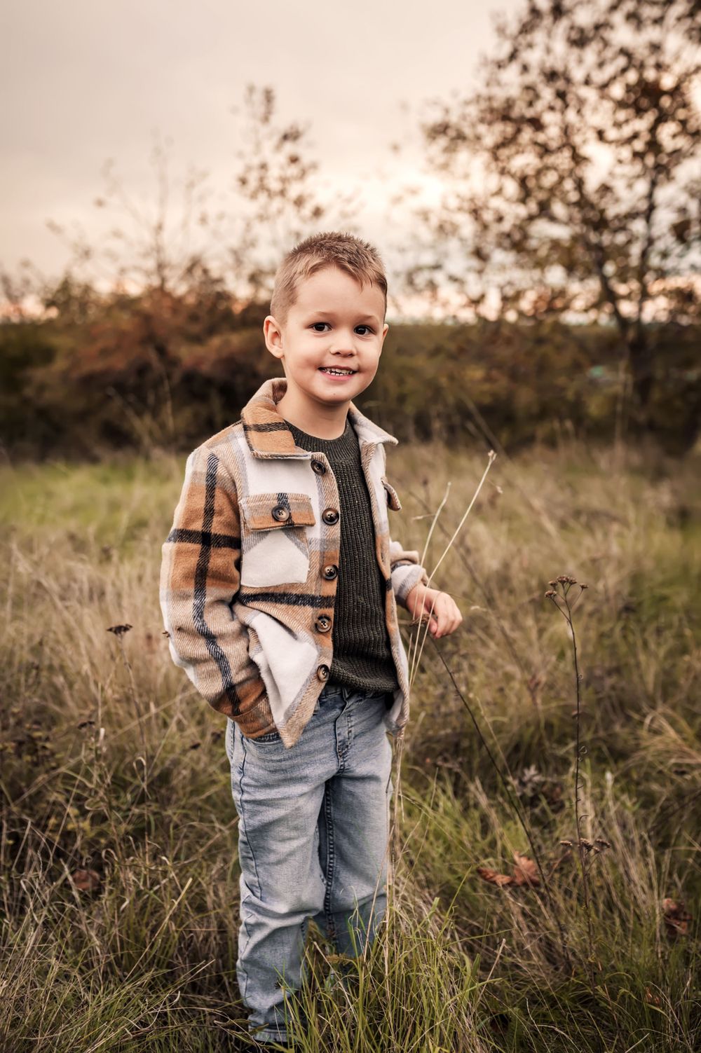 Young child in plaid jacket and jeans stands in tall grass during golden hour sunset in autumn field.