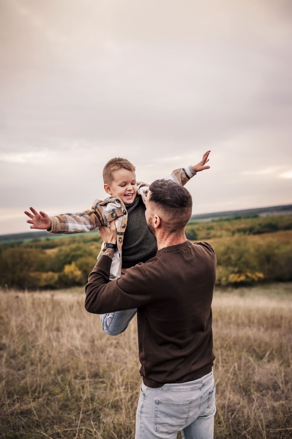 A small child wearing plaid is lifted playfully into the air against an autumn countryside backdrop at sunset.