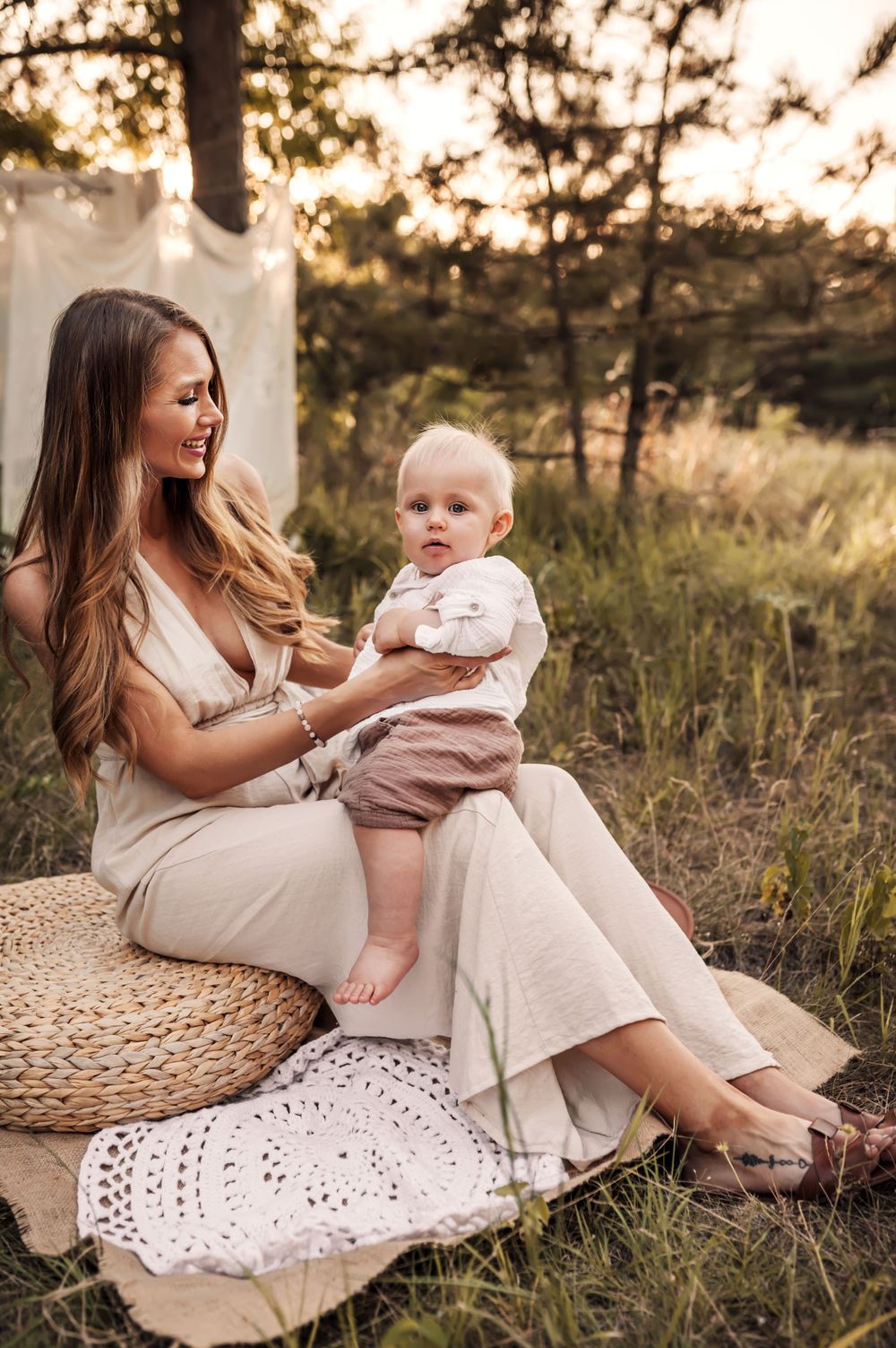 A peaceful outdoor portrait at sunset with a woman in beige dress seated on cushions while holding a baby in pink.