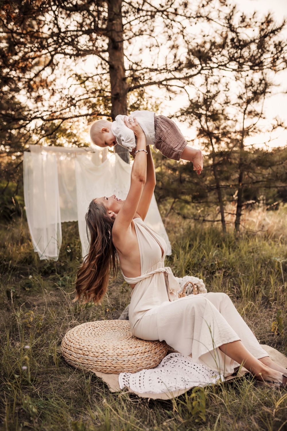 Two figures in white clothing share a tender moment on woven mat outdoors during golden sunset among pine trees.