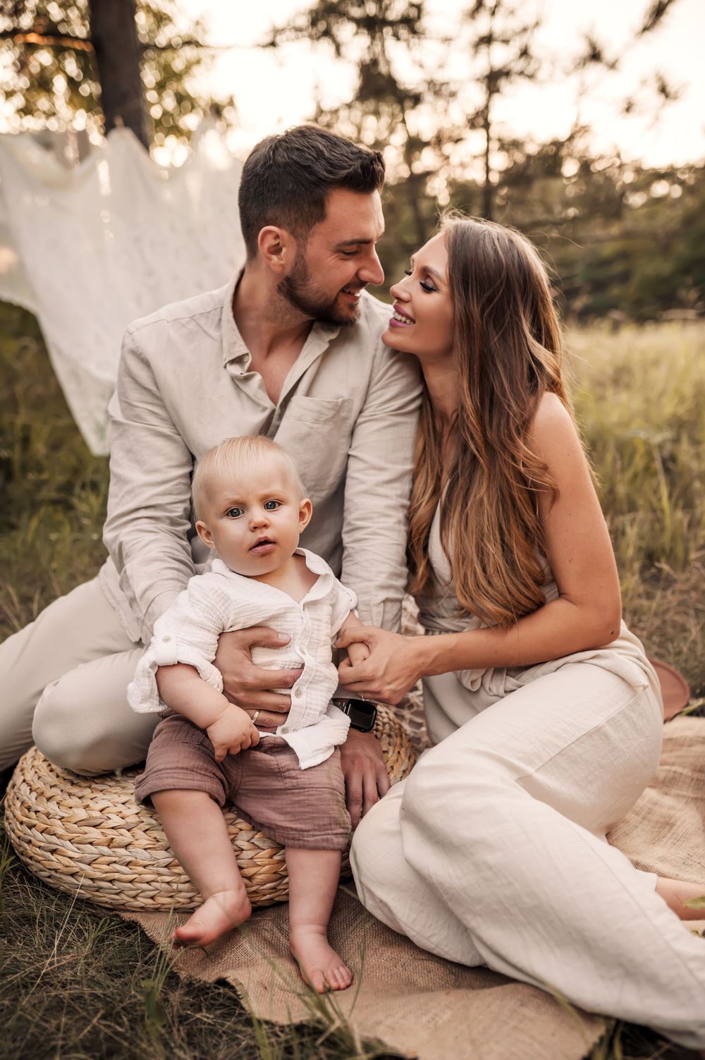A family in neutral outfits shares an intimate moment while sitting on a woven mat during golden hour sunset.