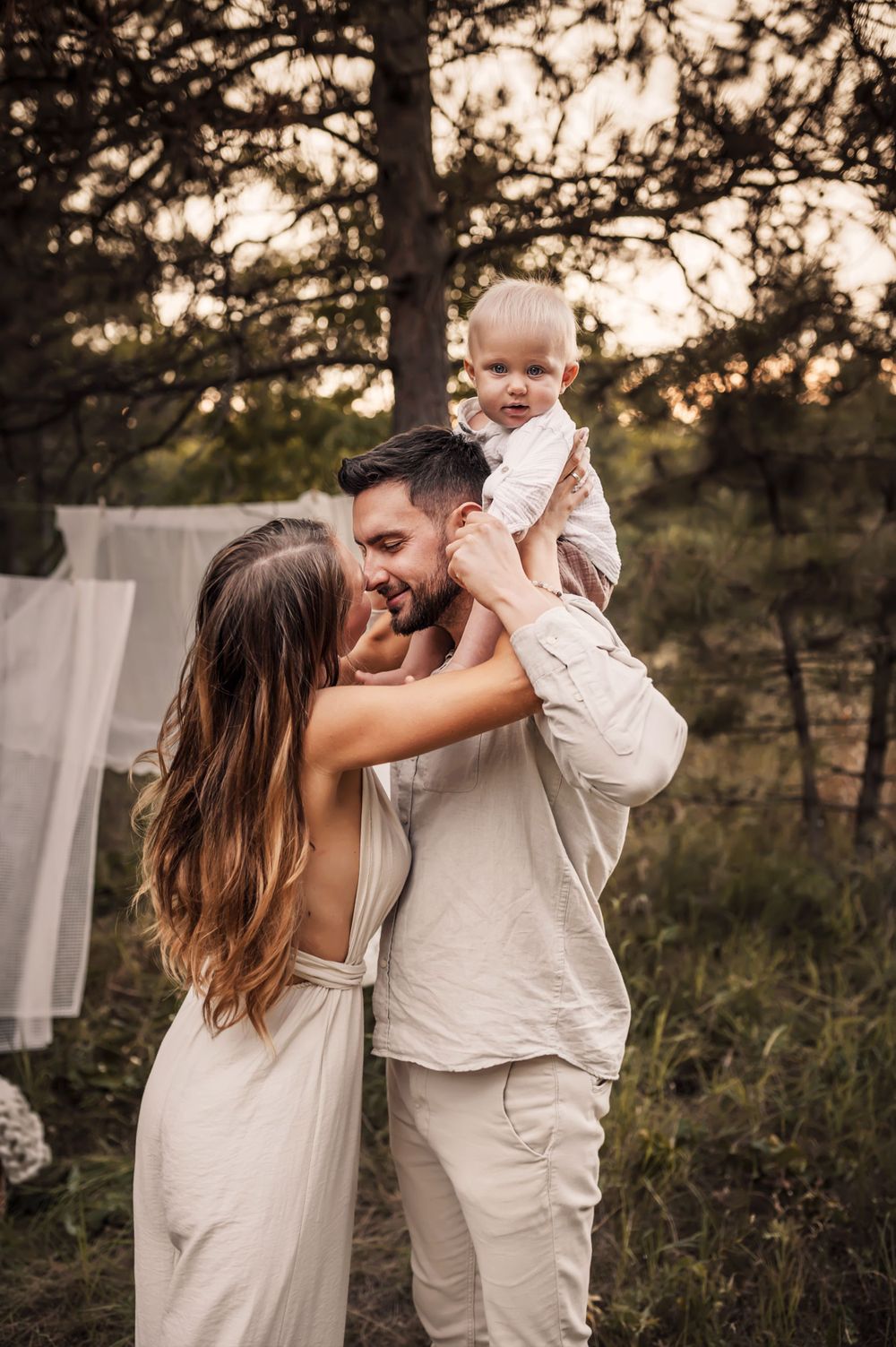 Family enjoying tender moment together outdoors with hanging linens at sunset in rustic woodland setting.