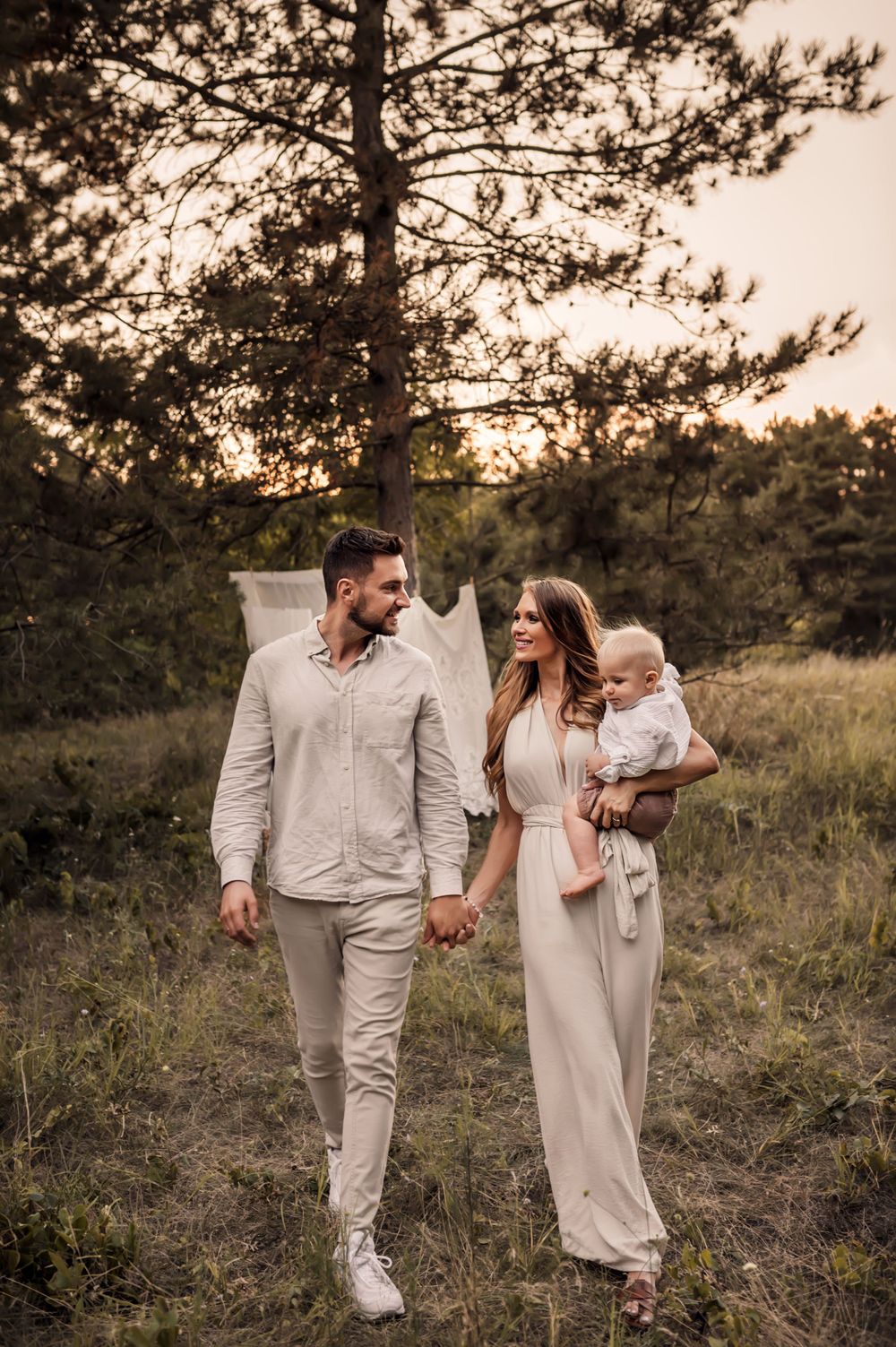 A couple in coordinated beige outfits walking together with their baby through a pine forest at sunset.