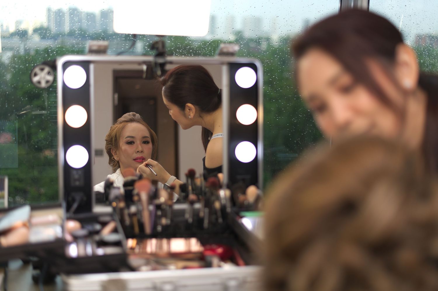 A reflection in a makeup mirror shows someone getting ready with cosmetics and lights.