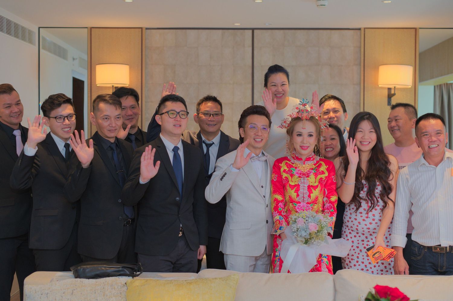 A group photo showing wedding guests in formal attire posing together with waves and smiles in an indoor setting.