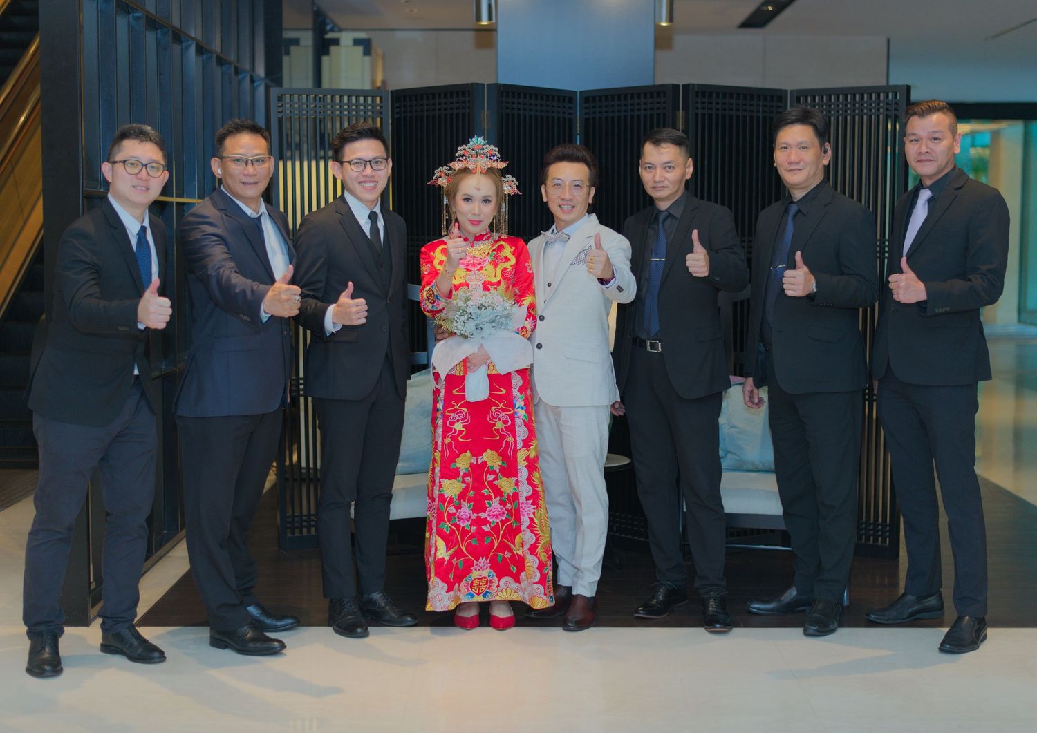 A wedding party lineup showing groomsmen in dark suits giving thumbs up beside a couple in traditional attire.