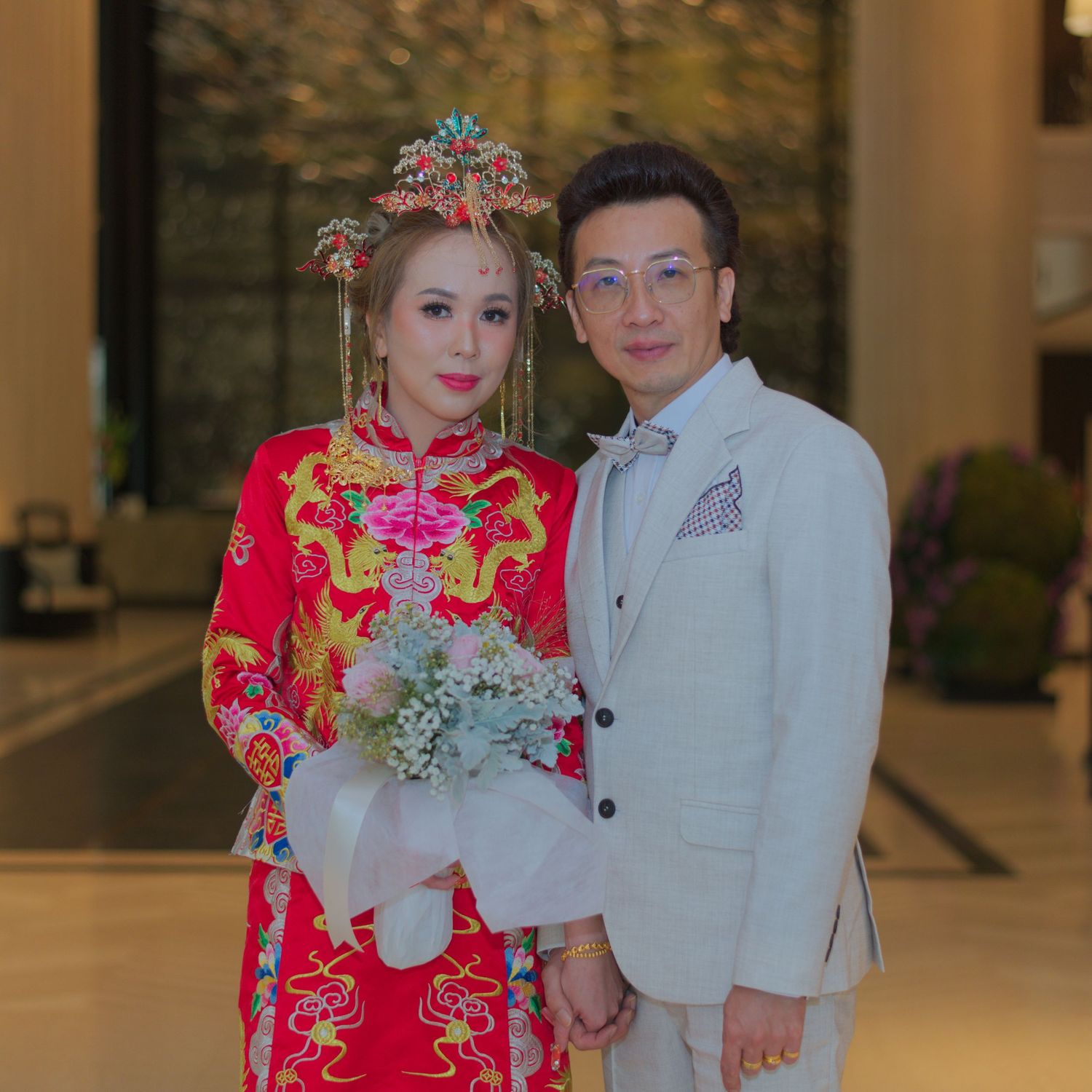 A bride in red traditional Chinese wedding dress stands with groom in white suit against an elegant indoor backdrop.