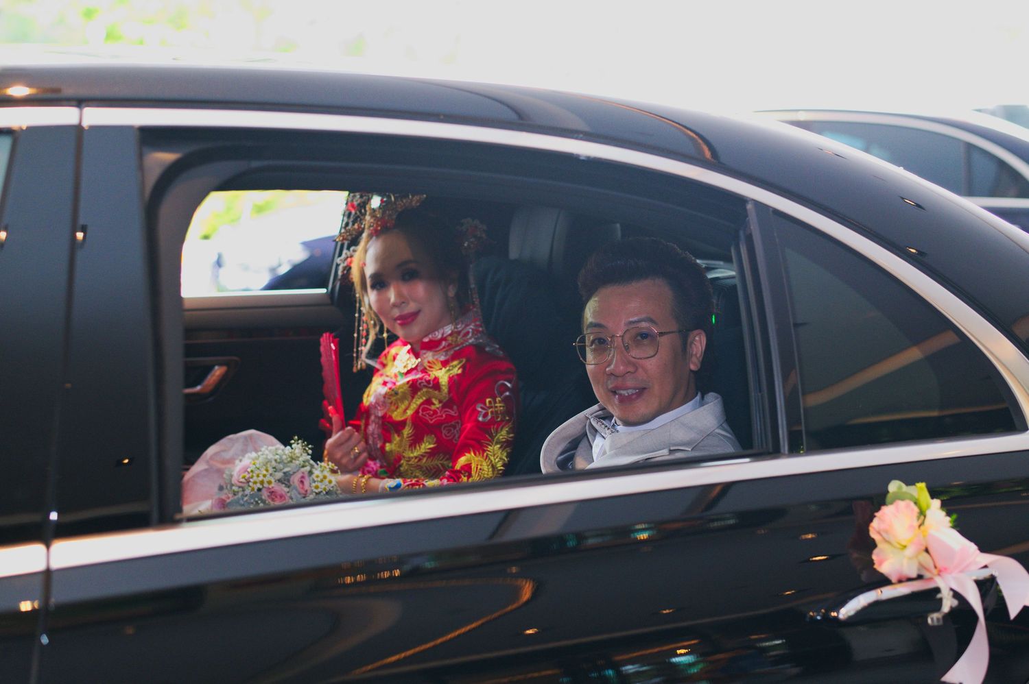 A couple in wedding attire sitting inside a black car during their celebration.