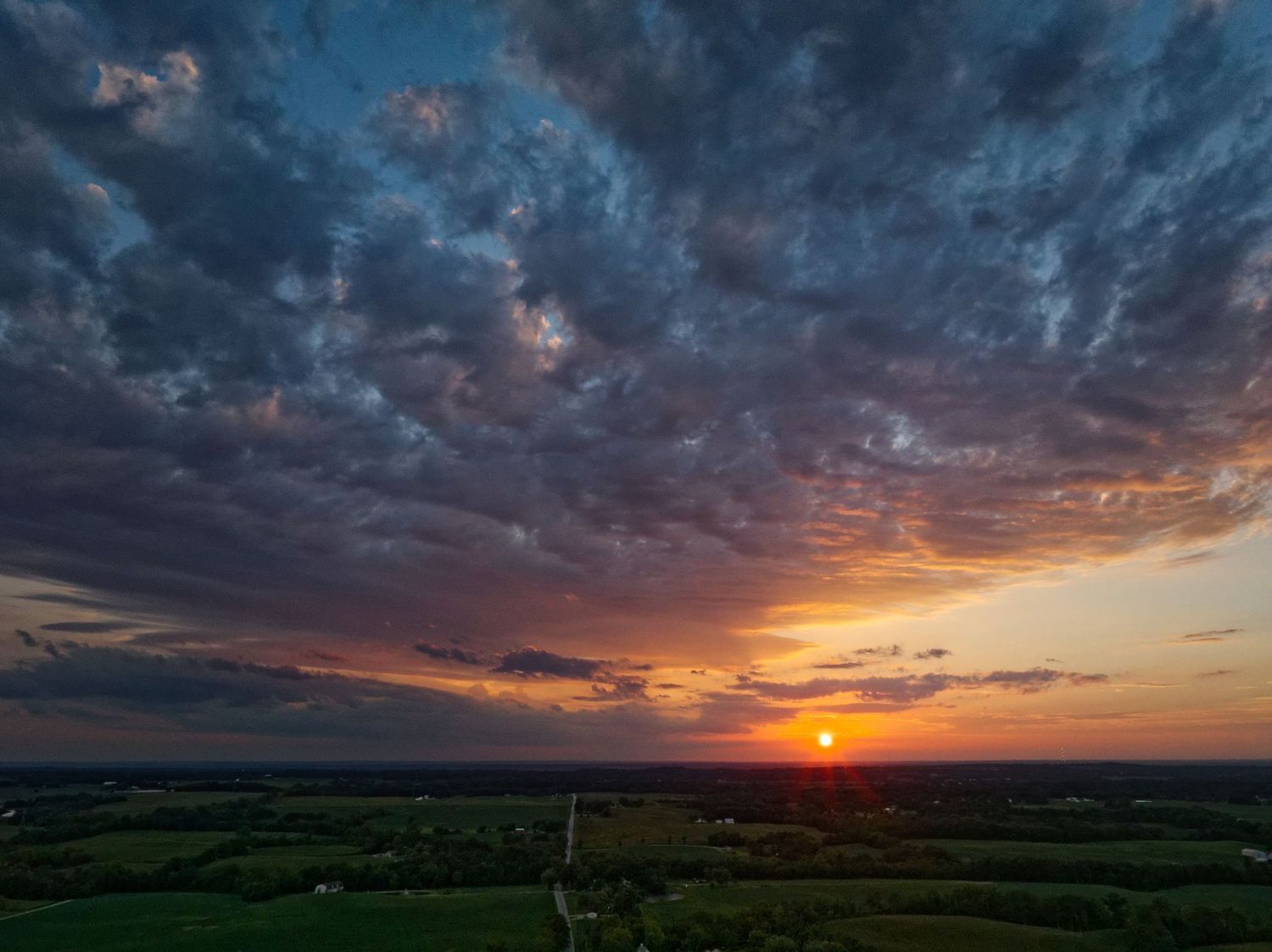 Aerial view of dramatic sunset over rural countryside with vibrant orange sun and deep blue storm clouds.