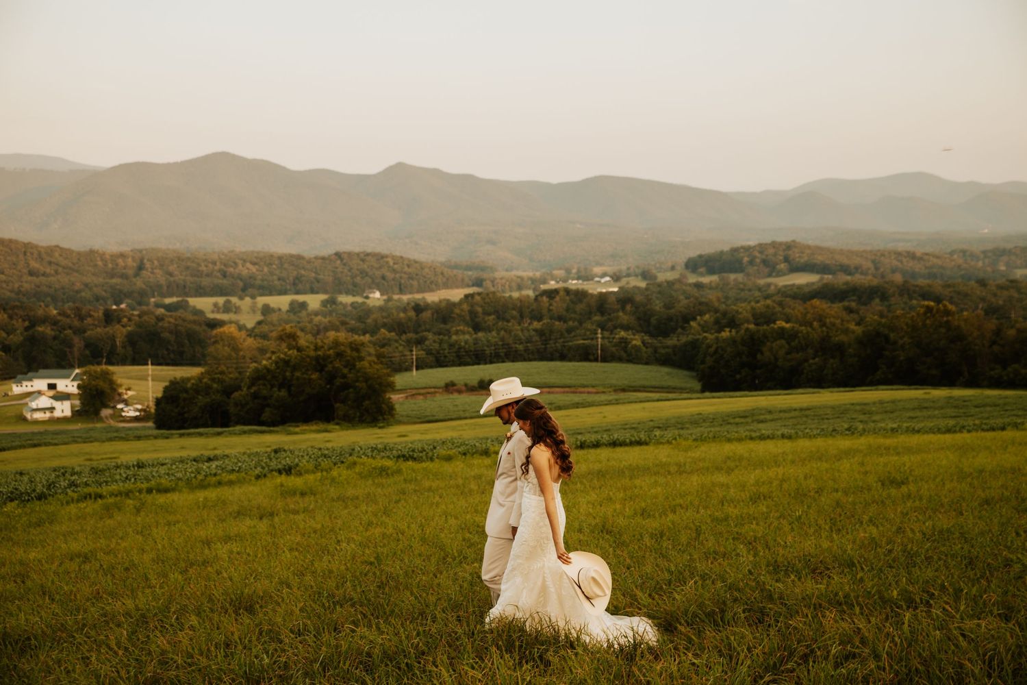 Bride in white gown and cowboy hat stands in lush green field with scenic mountain backdrop at golden hour.