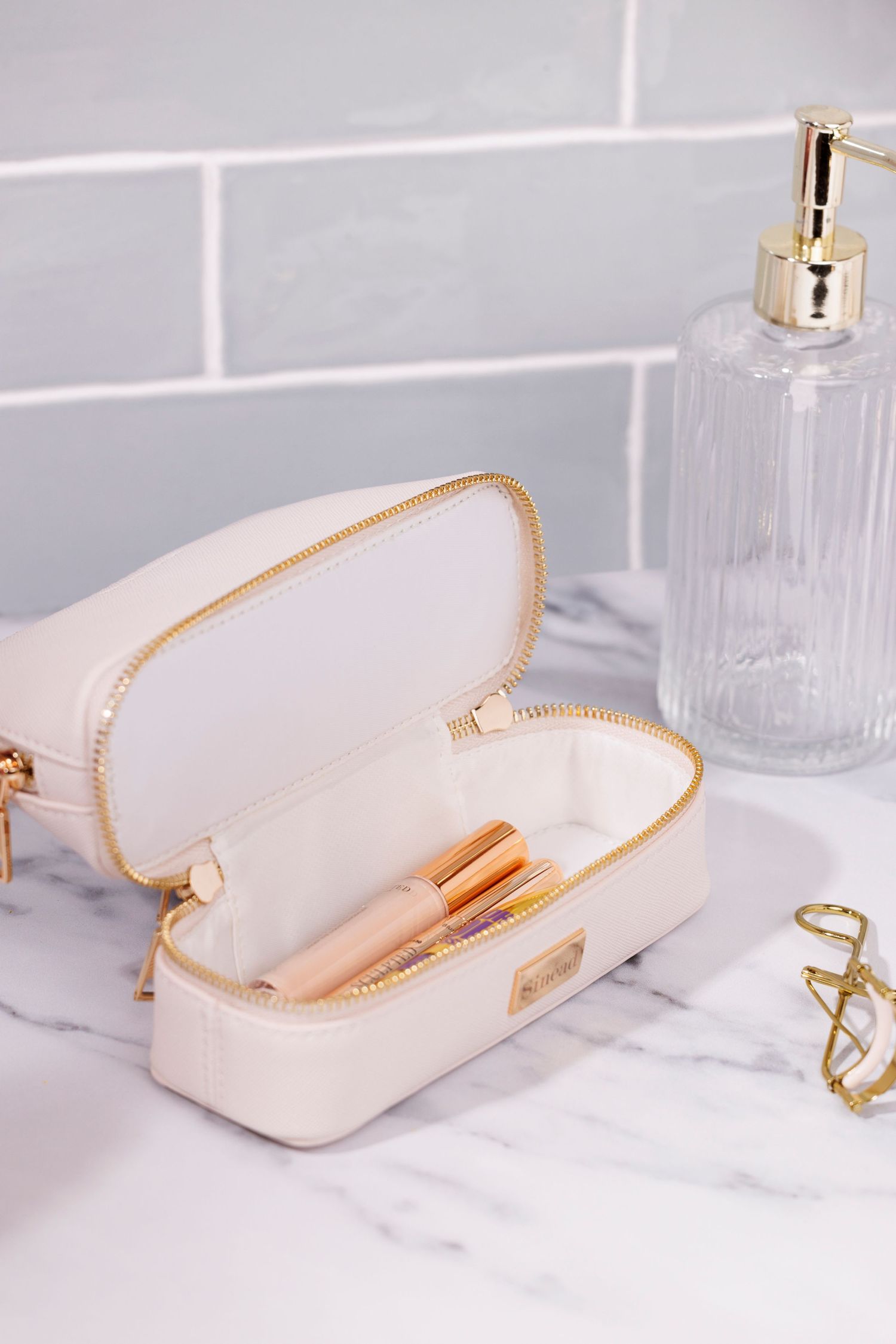 White cosmetic bag with gold trim open to display makeup brushes on marble counter and a tiled bathroom backdrop.