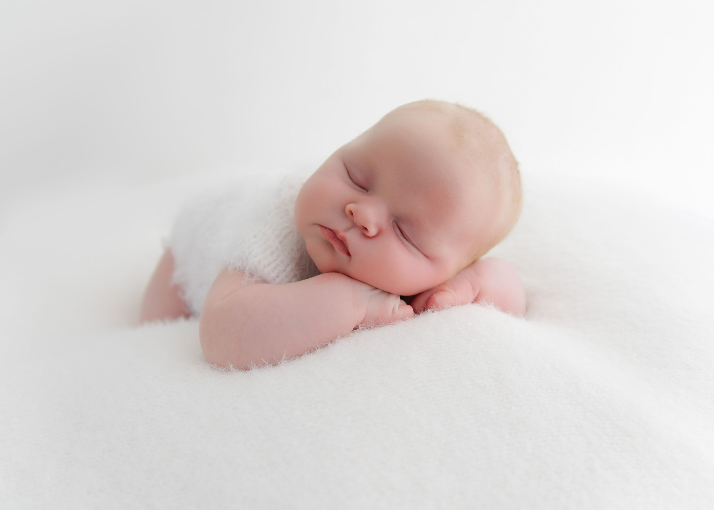 Peaceful newborn baby sleeping on a fluffy white blanket, wearing a soft white wrap, in a serene studio portrait.