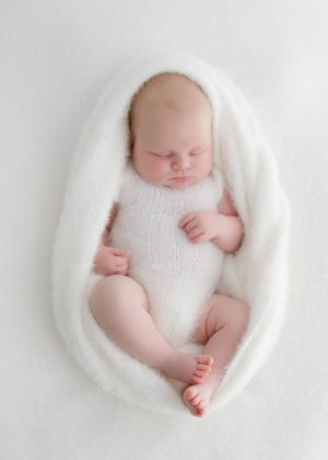 Sleeping newborn baby wrapped in soft white fluffy blanket wearing a white knit romper on a white background.