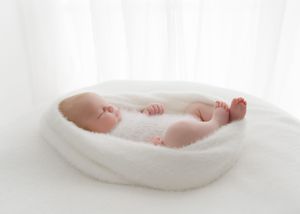 Newborn baby sleeping peacefully in a white fluffy bowl on a soft white blanket in a bright studio.