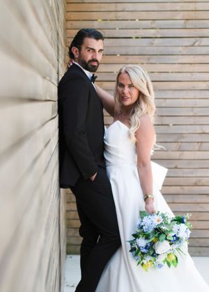 Bride in white gown holding blue bouquet leans with groom in black tuxedo against wooden wall.