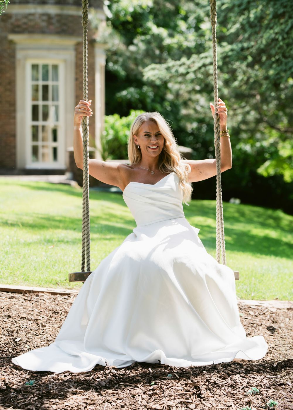 Bride in white strapless ball gown sitting on a rope swing outdoors, smiling joyfully on her wedding day.