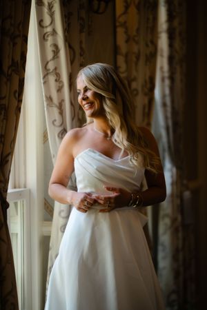 Smiling bride in strapless white wedding gown standing by a sunlit window with elegant curtains.