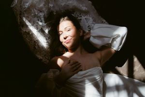 Woman lying down wrapped in white fabric, bathed in dramatic sunlight with a soft smile.