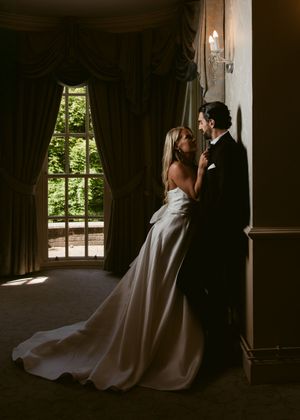 Bride in elegant white gown and groom in tuxedo share an intimate moment by a sunlit window in a grand estate.