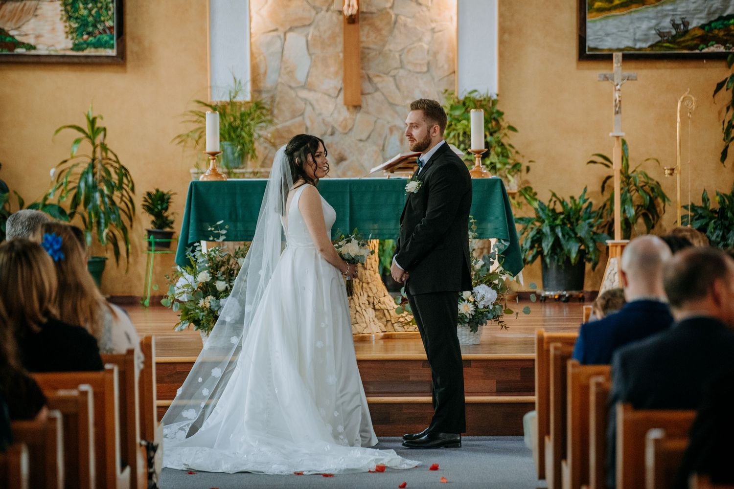 Couple exchanging vows at altar in church decorated with greenery and flowers.