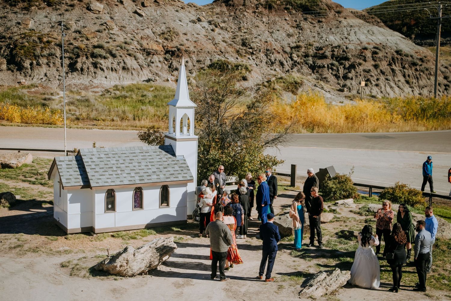 Small white chapel surrounded by rocky cliffs with a group of people gathered outside.