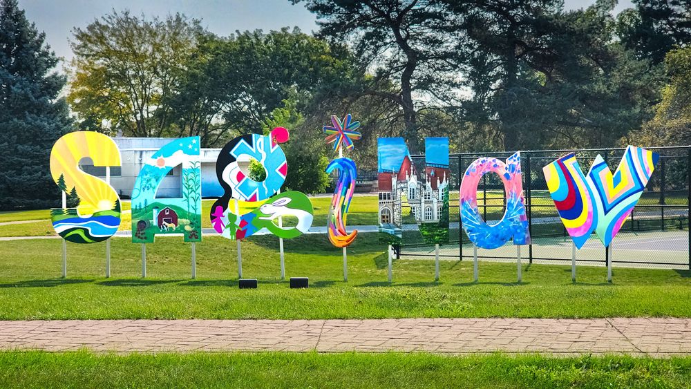 Colorful outdoor art installation spells out SAGINAW in large decorative letters on a grassy lawn surrounded by trees.