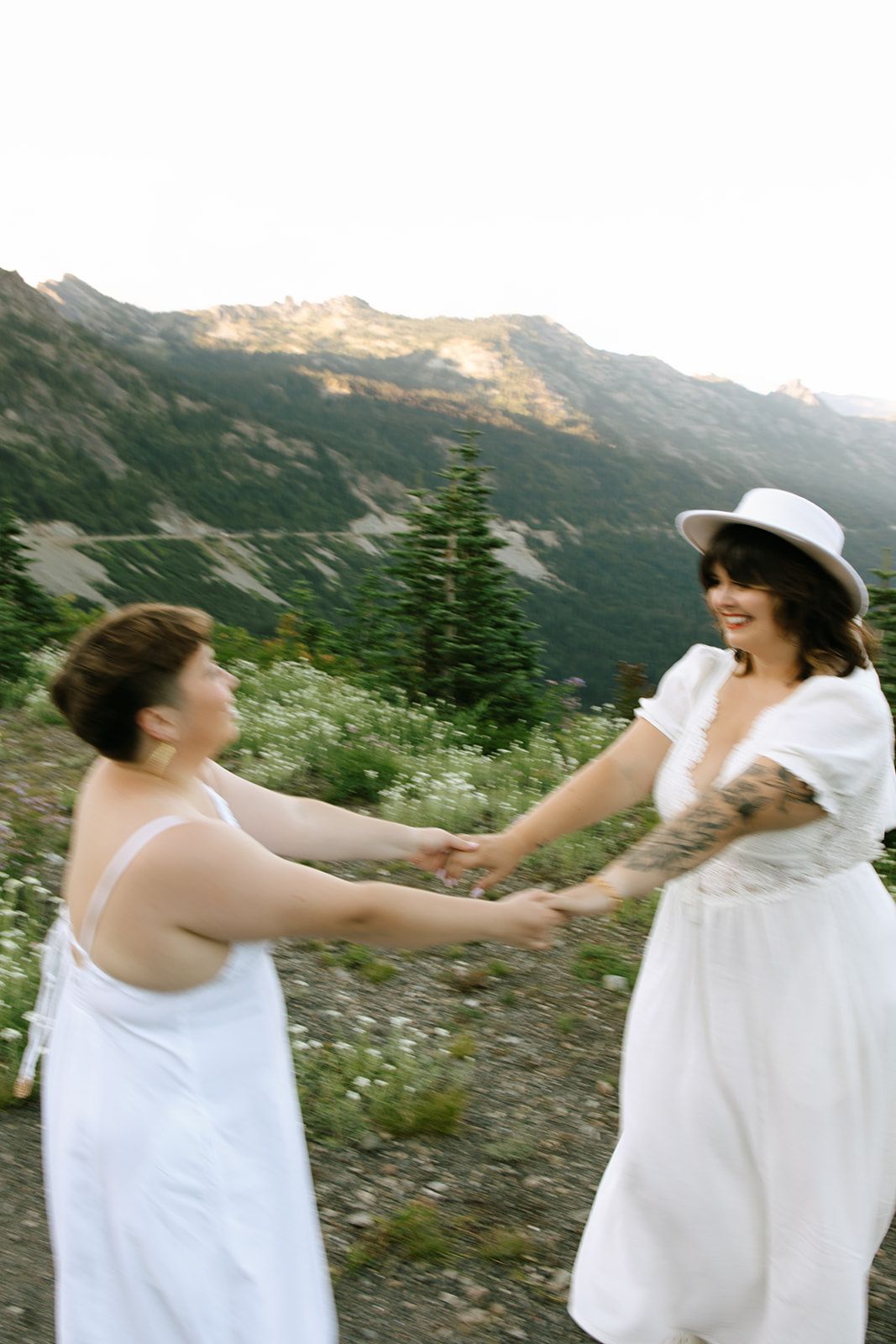 Two brides in white dresses hold hands and dance together on mountain wildflower meadow path.