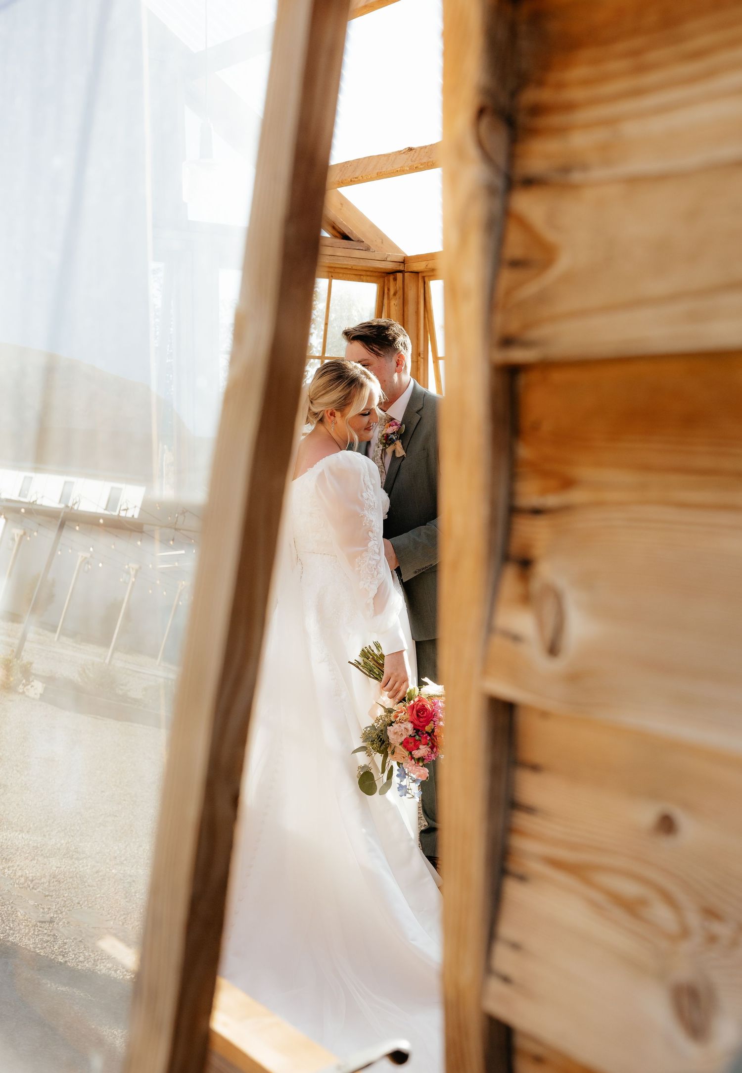 Bride and groom share a tender moment framed by rustic wooden walls, bride holding a pink bouquet.
