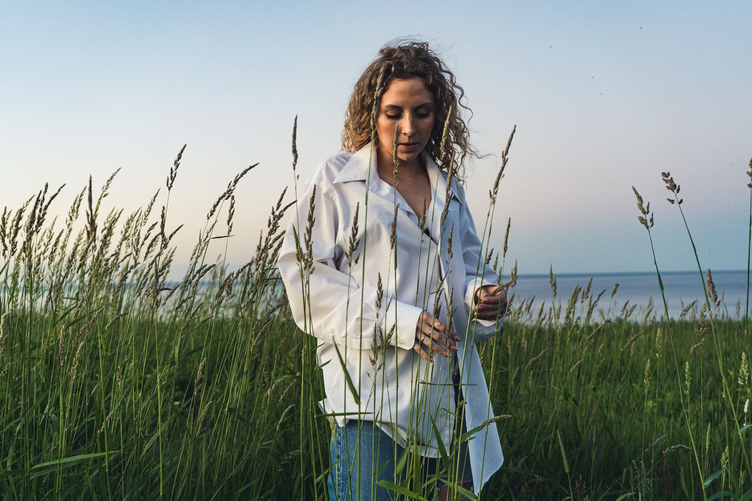 Person in white jacket walks through tall coastal grass near ocean at sunset.