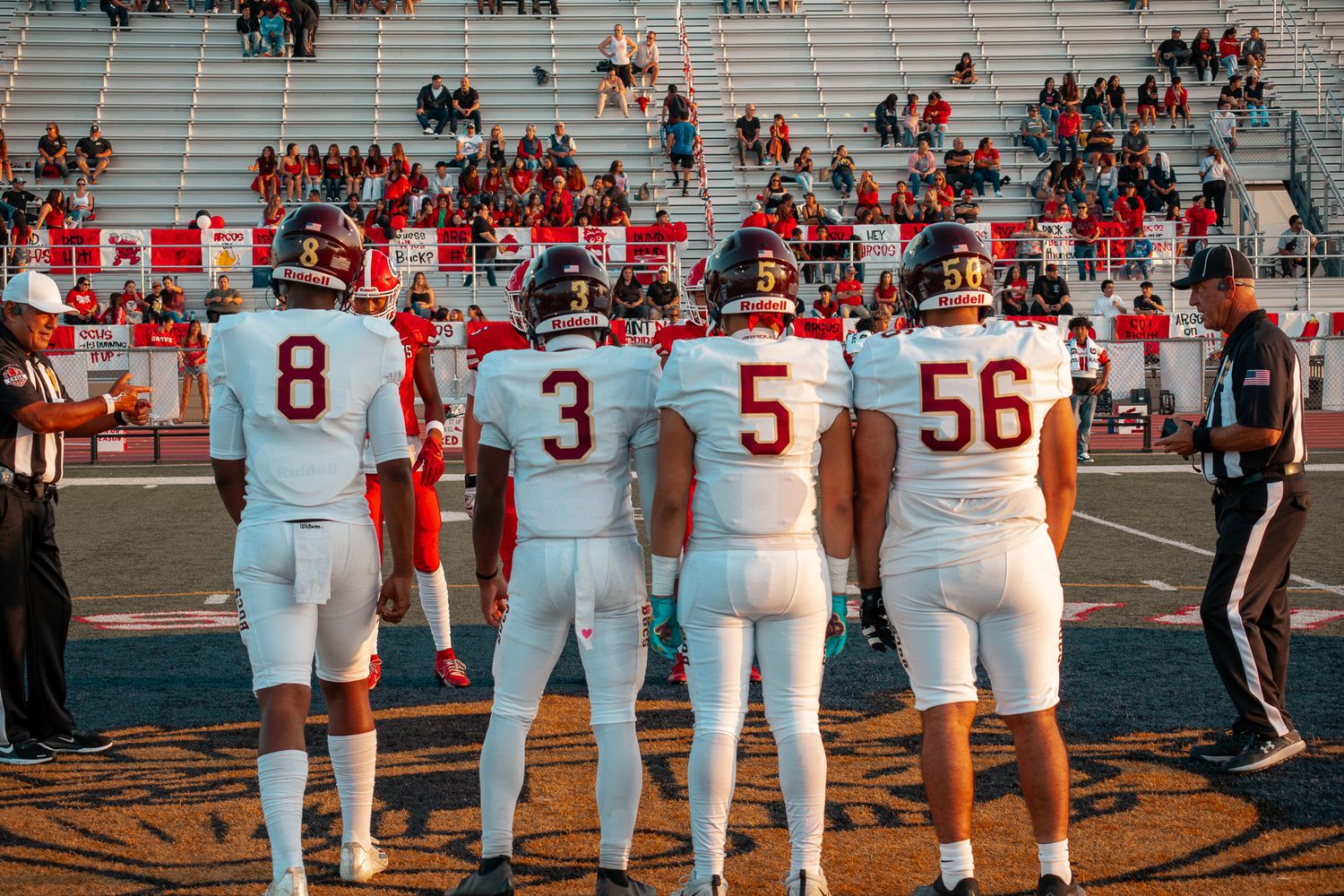 Row of football players lined up on sideline showing jersey numbers and uniforms from behind.