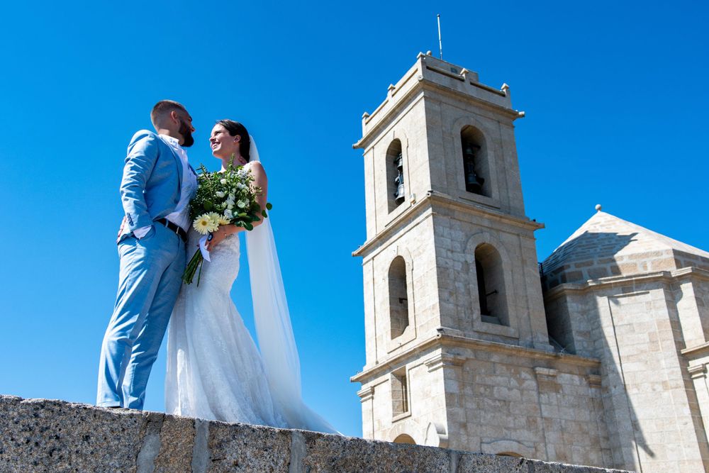 Wedding couple posing against white church tower and bright blue sky backdrop.