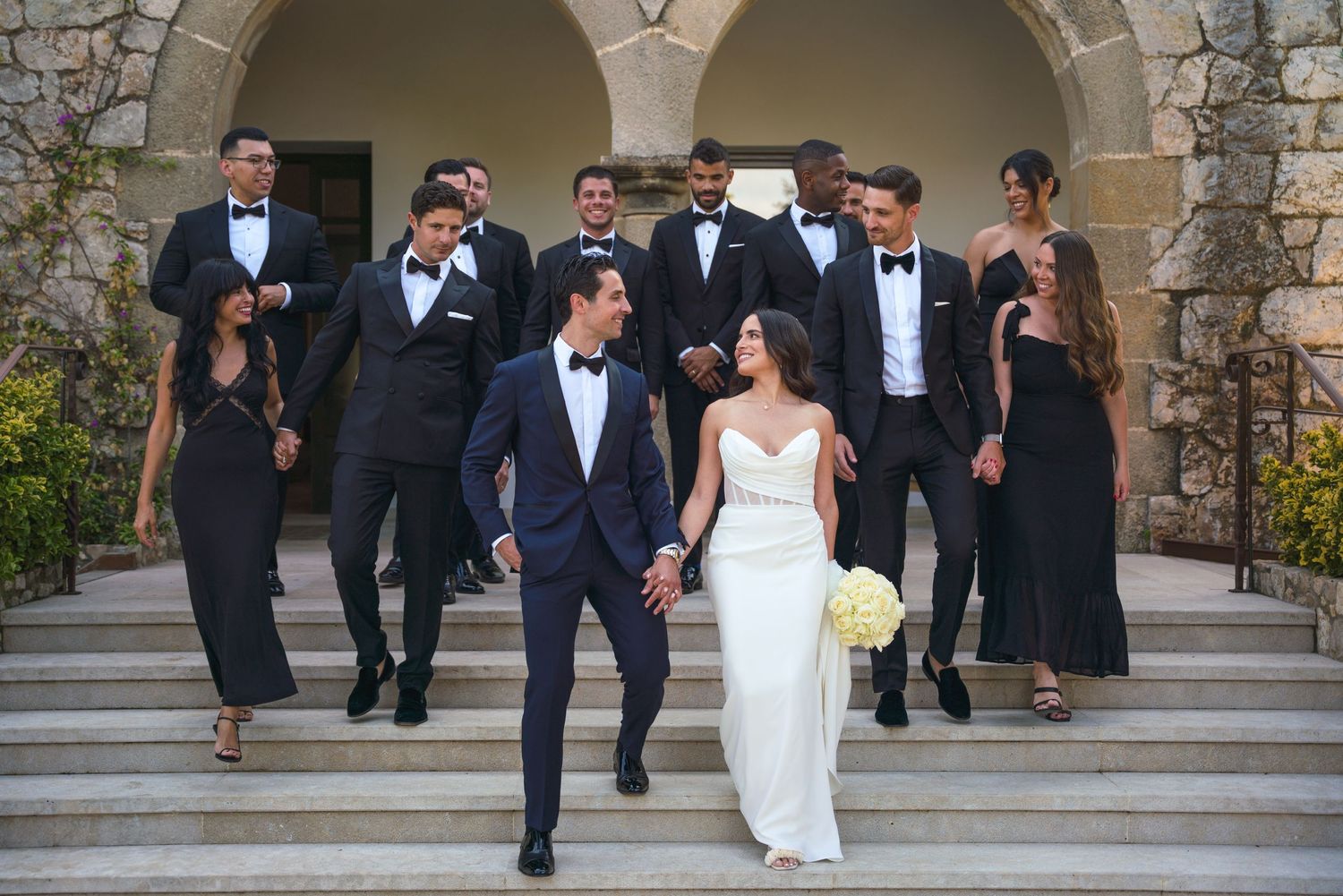 Wedding party in formal attire descends stone steps with bride and groom in the center.
