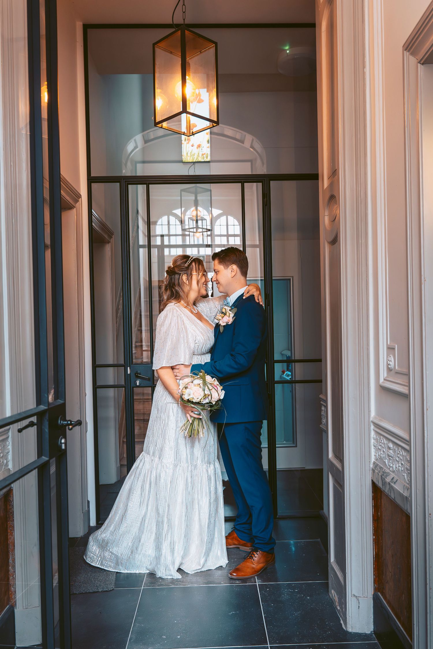 A tender wedding moment captured in an elegant hallway with hanging lantern and architectural details.