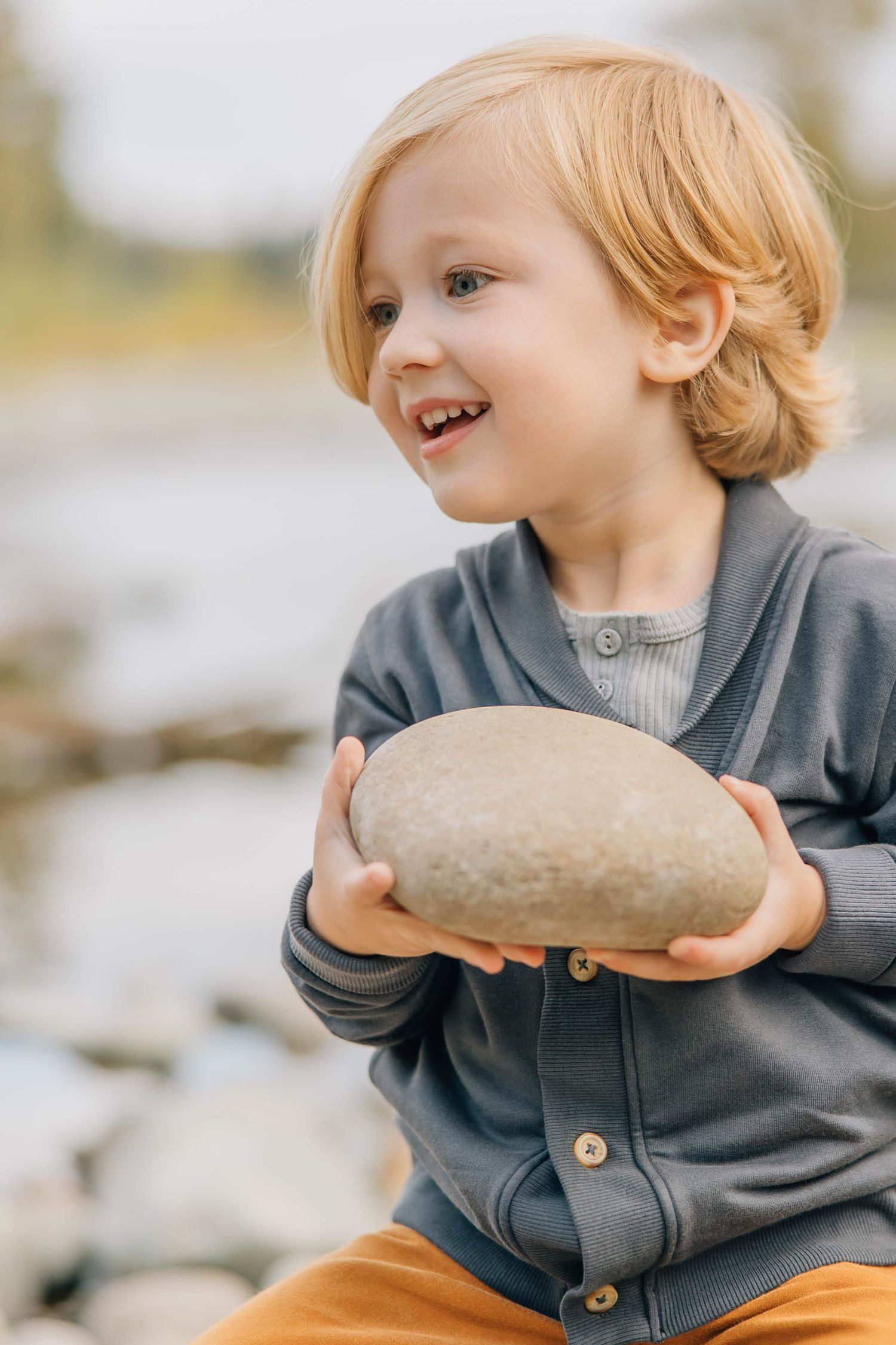 Adorable blonde toddler boy smiling while holding a large smooth rock near a river.