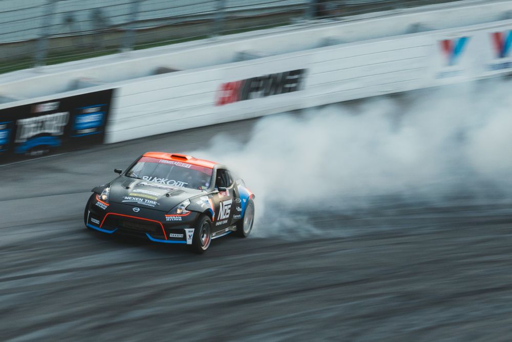 A black drift car slides sideways creating smoke on a race track.
