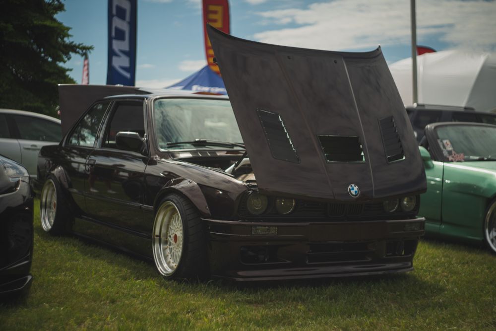 Modified black BMW E30 with hood up displaying engine bay at car show.